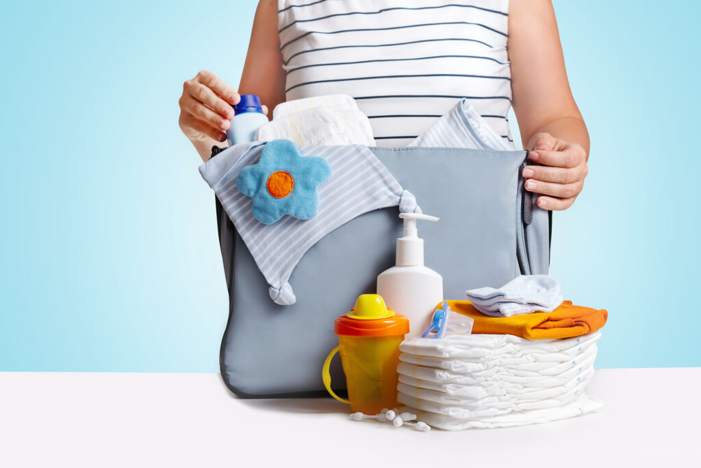 Unrecognizible pregnant caucasian woman in striped t-shirt packing big blue diaper bag to maternity hospital. Diapers, nappy, hat, bottle and other necessary things for newborn baby. Blue background.